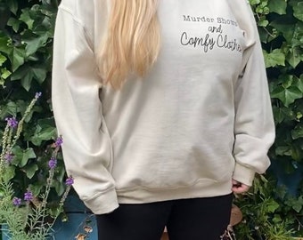 May include: A woman wearing a cream colored sweatshirt with the text "Murder Shows and Comfy Clothes" printed on the front. She is wearing black leggings and gray sneakers.