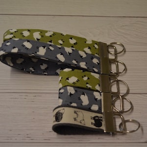 May include: Four keychains featuring different sheep designs. The keychains are made of fabric with a green, grey, and white pattern. The keychains are attached to a silver keyring.