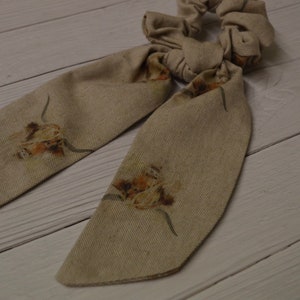 May include: A beige fabric hair scrunchie with a bow and floral print. The scrunchie has two long tails with a floral print.