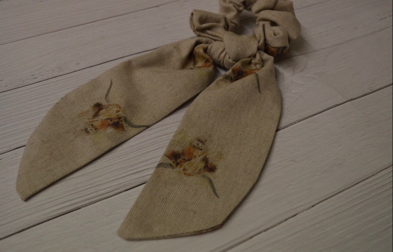 May include: A beige fabric hair scrunchie with a bow and floral print. The scrunchie has two long tails with a floral print.