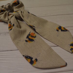 May include: A beige fabric hair scrunchie with a printed pattern of yellow and black bumblebees.