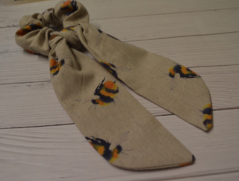 May include: A beige fabric hair scrunchie with a printed pattern of yellow and black bumblebees.
