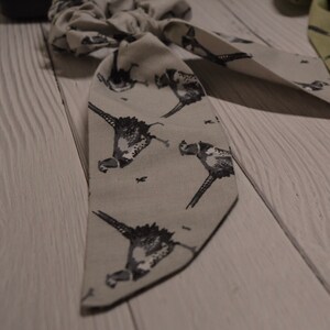May include: A gray fabric hair scrunchie with a pheasant print. The scrunchie is tied in a bow and has a long tail.