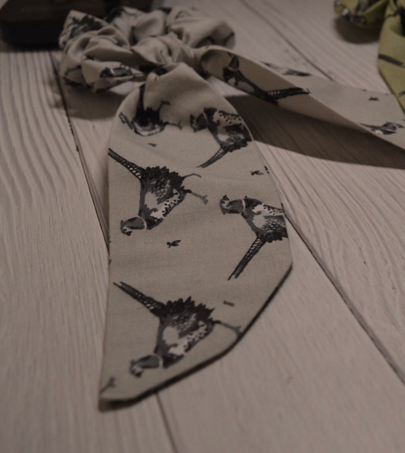 May include: A gray fabric hair scrunchie with a pheasant print. The scrunchie is tied in a bow and has a long tail.