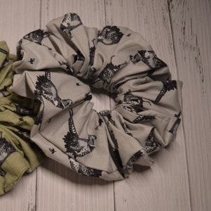 May include: A gray and white scrunchie with a black and white bird print. The scrunchie is made of fabric and is gathered into a circle.