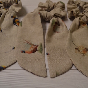 May include: Three beige fabric hair scrunchies with long tails. The tails have a repeating pattern of small birds and flowers.
