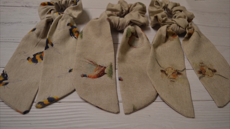 May include: Three beige fabric hair scrunchies with long tails. The tails have a repeating pattern of small birds and flowers.