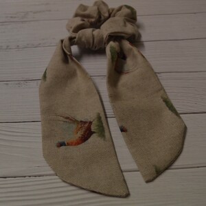 May include: A beige fabric hair scrunchie with a pheasant print. The scrunchie has two long tails that are tied together.