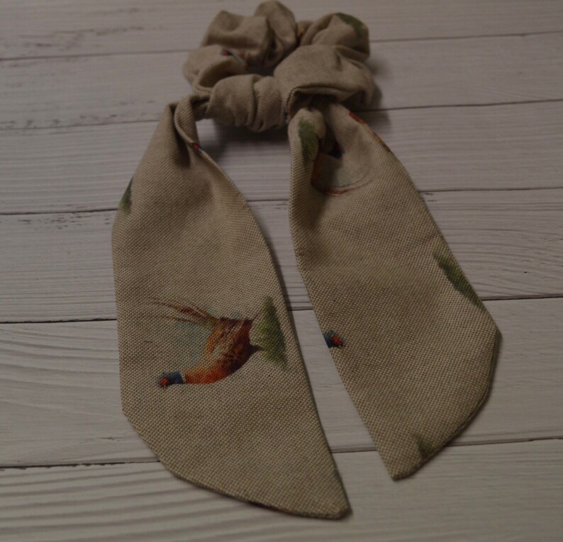 May include: A beige fabric hair scrunchie with a pheasant print. The scrunchie has two long tails that are tied together.