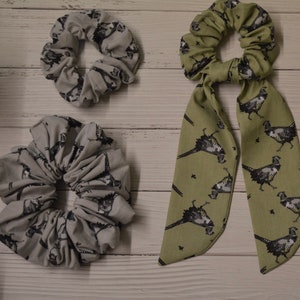 May include: Two hair scrunchies with a pheasant print. One is a gray scrunchie and the other is a green scrunchie with a long bow.
