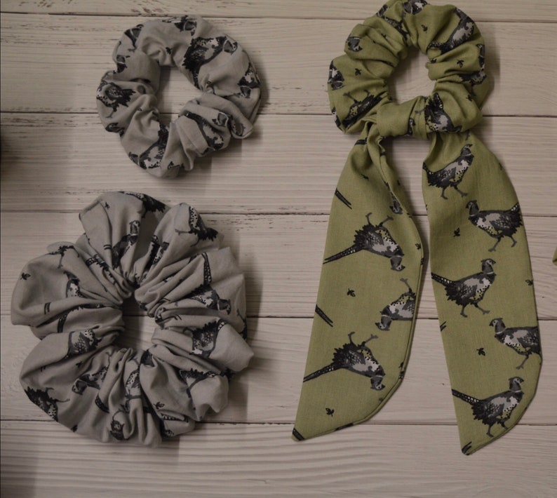 May include: Two hair scrunchies with a pheasant print. One is a gray scrunchie and the other is a green scrunchie with a long bow.