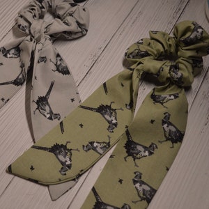 May include: Two fabric hair scrunchies with matching fabric ties. One scrunchie is green with a pheasant print and the other is gray with a pheasant print.