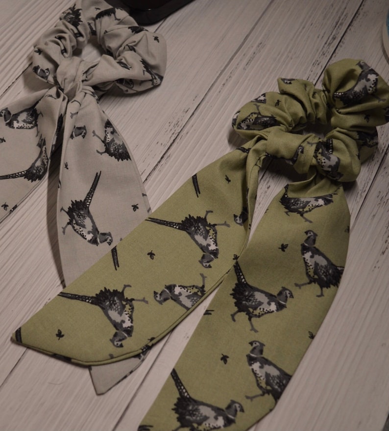 May include: Two fabric hair scrunchies with matching fabric ties. One scrunchie is green with a pheasant print and the other is gray with a pheasant print.
