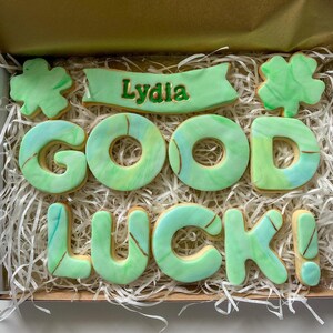 Puede incluir: Una caja de galletas decorada en verde y dorado con las palabras "Lydia Good Luck!" y un dise&ntilde;o de tr&eacute;bol de cuatro hojas.