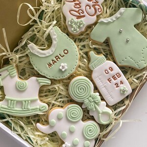 May include: A collection of baby-themed cookies in a gift box. The cookies are decorated with white and light green icing, featuring designs like a bib with the name "MARLO", a baby bottle, a onesie, a rattle, a rocking horse, and an elephant. The cookies are arranged on a bed of shredded paper.