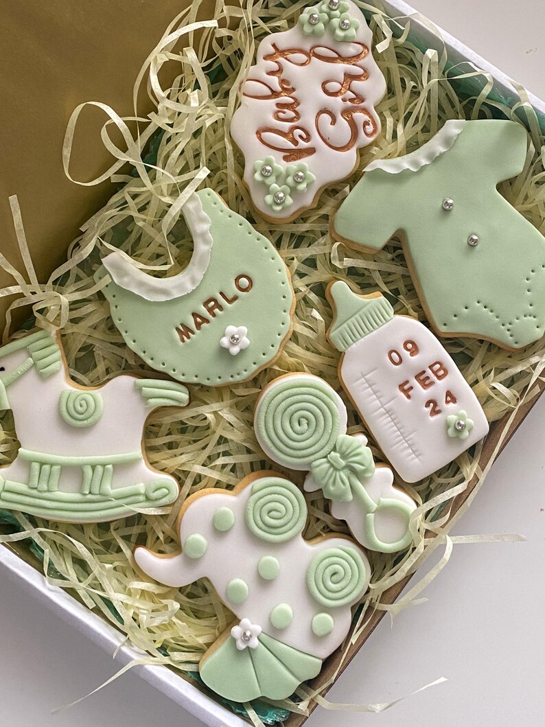 May include: A collection of baby-themed cookies in a gift box. The cookies are decorated with white and light green icing, featuring designs like a bib with the name "MARLO", a baby bottle, a onesie, a rattle, a rocking horse, and an elephant. The cookies are arranged on a bed of shredded paper.