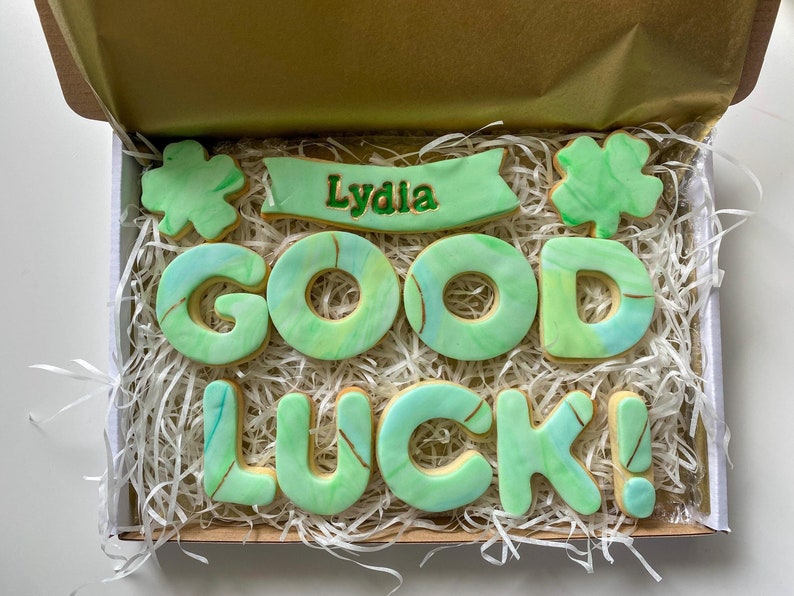 Puede incluir: Una caja de galletas decorada en verde y dorado con las palabras "Lydia Good Luck!" y un dise&ntilde;o de tr&eacute;bol de cuatro hojas.