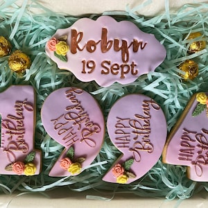May include: A set of six pink and purple cookies decorated with gold lettering and floral accents. The cookies spell out "Happy Birthday" and include the name "Robyn" and the date "19 Sept".