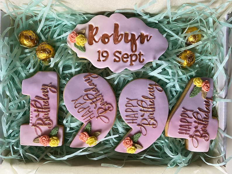 May include: A set of six pink and purple cookies decorated with gold lettering and floral accents. The cookies spell out "Happy Birthday" and include the name "Robyn" and the date "19 Sept".