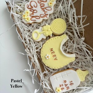 May include: A box of pastel yellow baby-themed cookies. The cookies include a cloud-shaped cookie with "Baby Girl" in gold script, a rattle, a bib with a name, a lollipop, and a baby bottle with a date.
