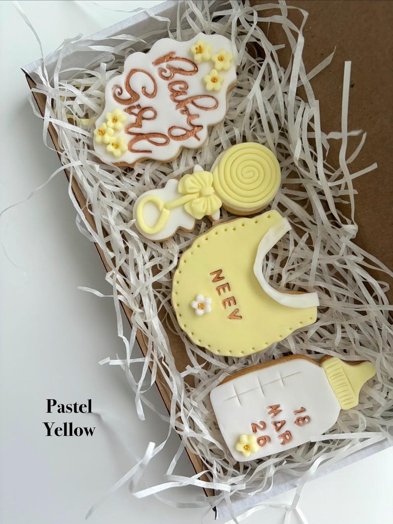 May include: A box of pastel yellow baby-themed cookies. The cookies include a cloud-shaped cookie with "Baby Girl" in gold script, a rattle, a bib with a name, a lollipop, and a baby bottle with a date.