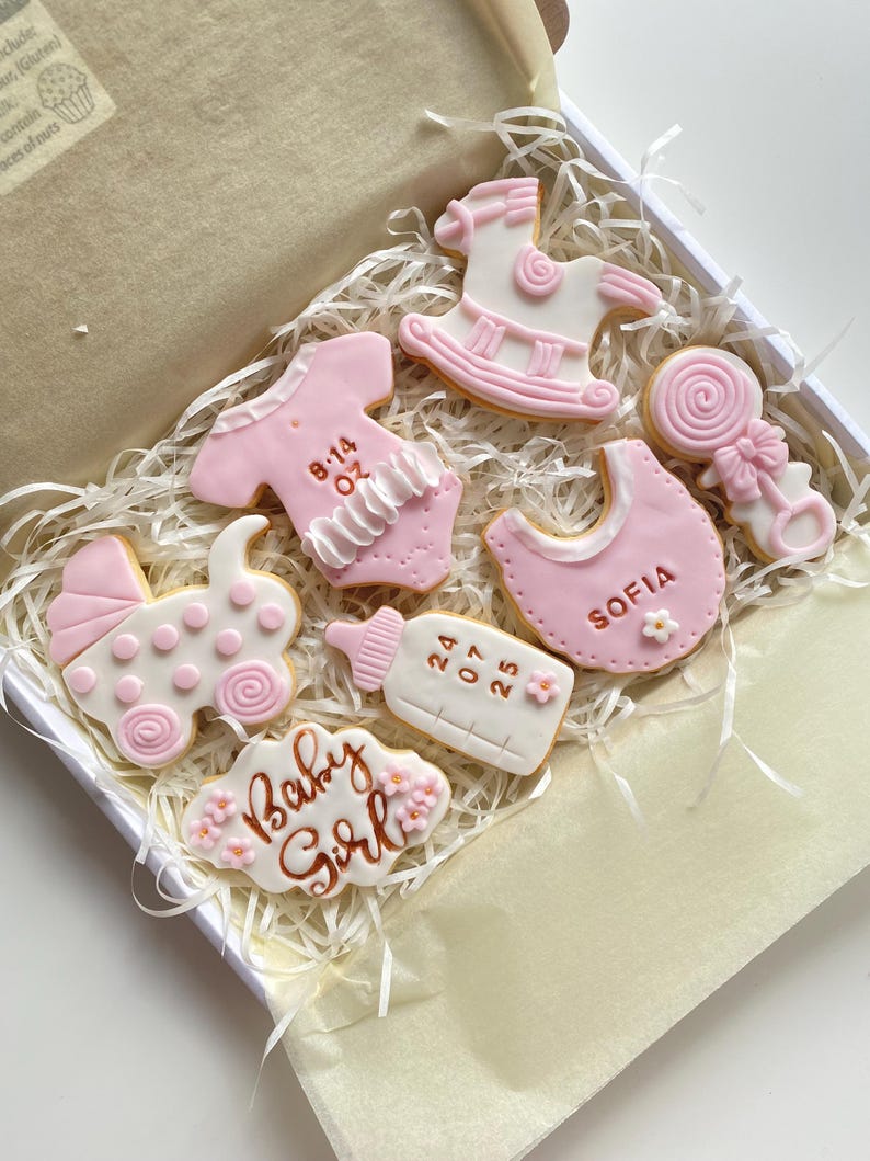 May include: A box of decorated baby-themed cookies. The cookies are in the shape of a baby carriage, bib, baby bottle, onesie, rocking horse, and a lollipop. The cookies are pink and white with floral accents and the words "Baby Girl".