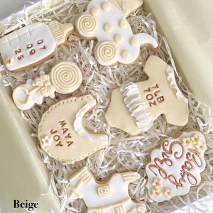May include: A box of beige and white baby-themed cookies. The cookies are shaped like a baby bottle, stroller, rattle, bib, onesie, rocking horse, and a cloud with the words "Baby Girl". Some cookies have dates and names.