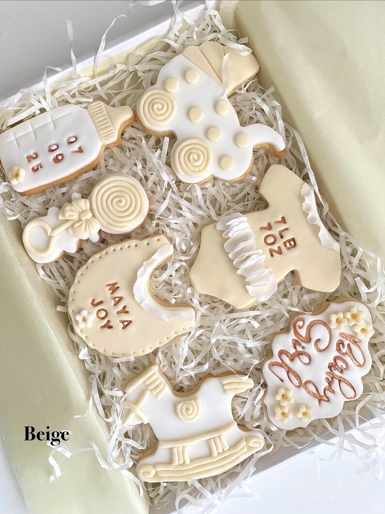 May include: A box of beige and white baby-themed cookies. The cookies are shaped like a baby bottle, stroller, rattle, bib, onesie, rocking horse, and a cloud with the words "Baby Girl". Some cookies have dates and names.