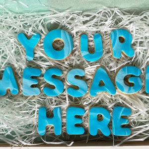 May include: A set of blue and white marbled cookies that spell out "YOUR MESSAGE HERE". The cookies are arranged in a box filled with white shredded paper.
