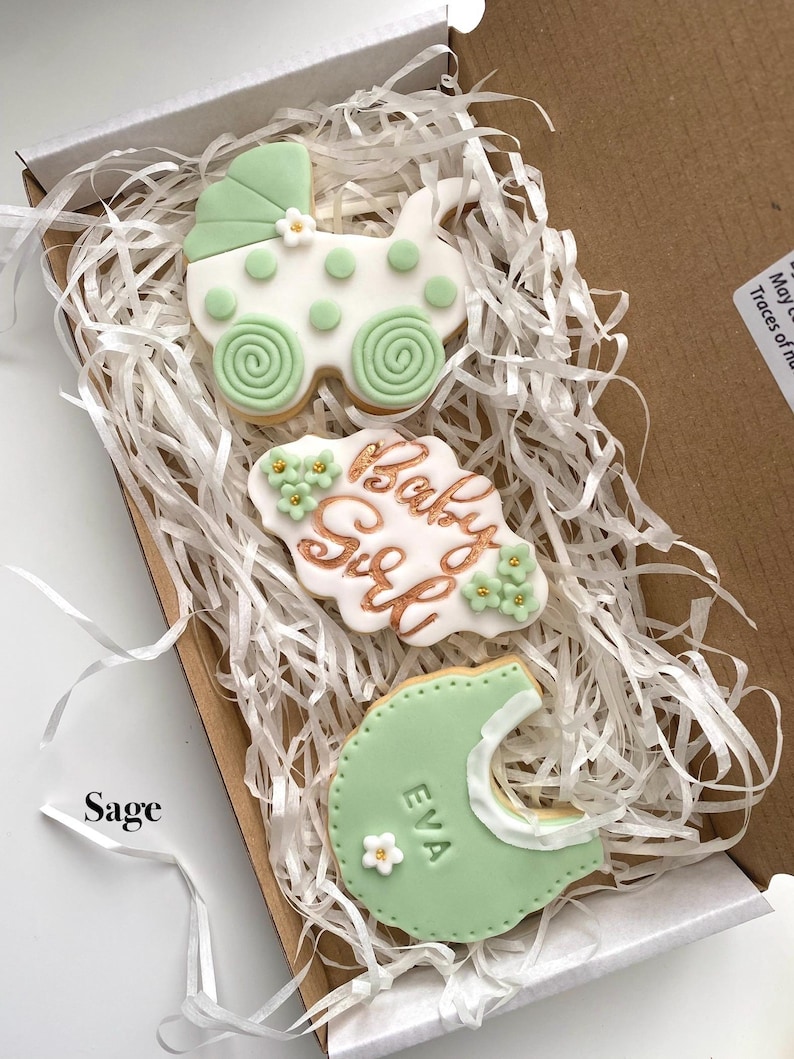 May include: A box of decorated sugar cookies, including a baby carriage, a plaque with "Baby Girl" in gold lettering, and a bib with the name "EVA". The cookies are white and sage green, with floral accents.
