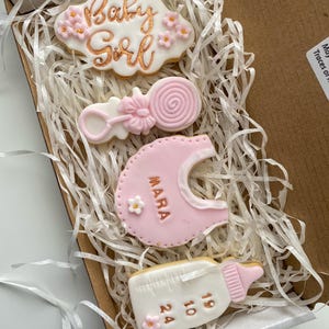May include: A box of decorated sugar cookies for a baby girl. The cookies include a cloud with "Baby Girl" in gold script, a rattle, a bib with "Mara", and a baby bottle with numbers. The cookies are pink, white, and gold.