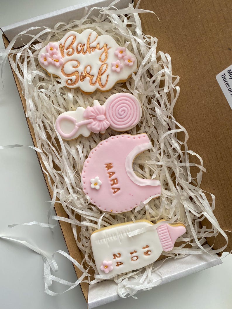 May include: A box of decorated sugar cookies for a baby girl. The cookies include a cloud with "Baby Girl" in gold script, a rattle, a bib with "Mara", and a baby bottle with numbers. The cookies are pink, white, and gold.
