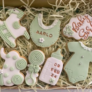 May include: A box of decorated baby-themed cookies. The cookies are white and light green, shaped like a rocking horse, bib, baby bottle, onesie, and a cookie that says "Baby Girl". The bib cookie has the name "Marlo" on it.