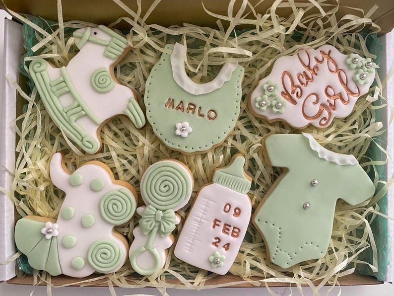 May include: A box of decorated baby-themed cookies. The cookies are white and light green, shaped like a rocking horse, bib, baby bottle, onesie, and a cookie that says "Baby Girl". The bib cookie has the name "Marlo" on it.