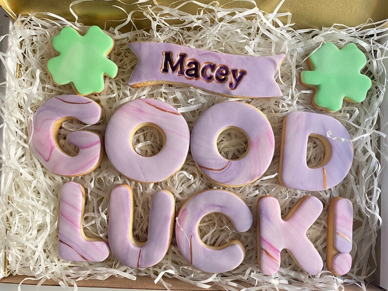 Puede incluir: Una caja de regalo llena de galletas marmoleadas de color p&uacute;rpura y dorado que deletrean "GOOD LUCK" con una galleta de tr&eacute;bol verde y una galleta de pancarta que dice "Macey".