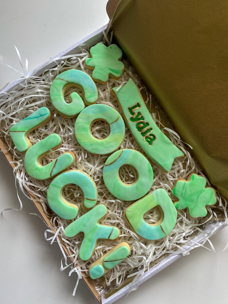 Puede incluir: Un conjunto de galletas "Buena suerte" verde y dorado en una caja blanca con papel triturado. Las galletas est&aacute;n decoradas con un dise&ntilde;o verde y dorado marmoleado. El conjunto incluye una pancarta con el nombre "Lydia" escrito en dorado.