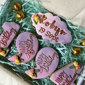 May include: A gift box filled with pink and purple cookies decorated with flowers and gold lettering. The cookies say "Happy Birthday" and "Robyn 19 Sept".