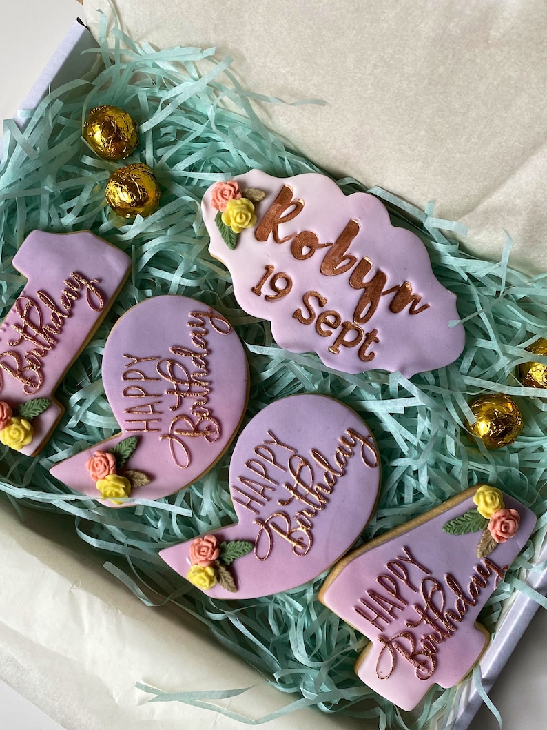 May include: A gift box filled with pink and purple cookies decorated with flowers and gold lettering. The cookies say "Happy Birthday" and "Robyn 19 Sept".