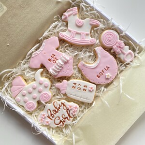 May include: A box of decorated sugar cookies in a baby shower theme. The cookies are white and pink, shaped like a baby carriage, bib, rocking horse, baby bottle, and onesie. Some cookies have text that reads "Baby Girl" and "Sofia".