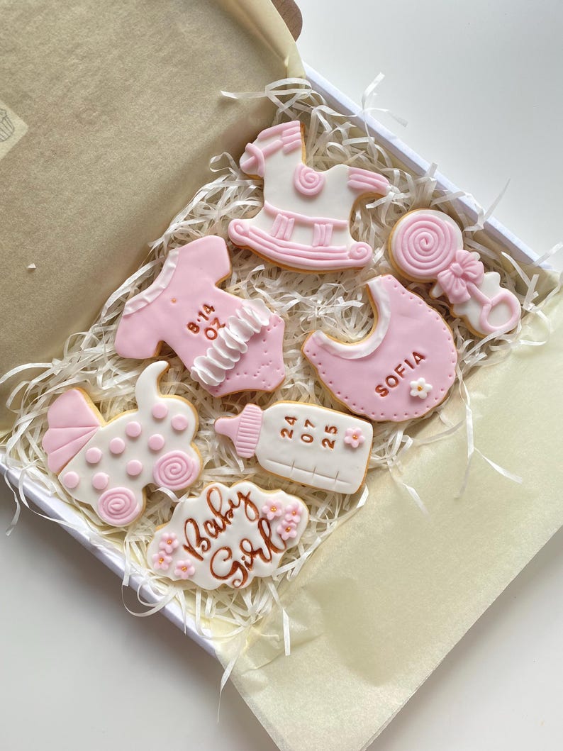 May include: A box of decorated sugar cookies in a baby shower theme. The cookies are white and pink, shaped like a baby carriage, bib, rocking horse, baby bottle, and onesie. Some cookies have text that reads "Baby Girl" and "Sofia".