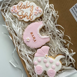 May include: A gift box filled with decorative cookies for a baby girl. The cookies include a cloud-shaped cookie with "Baby Girl" in gold script, a bib-shaped cookie, and a stroller-shaped cookie, all in pink and white.