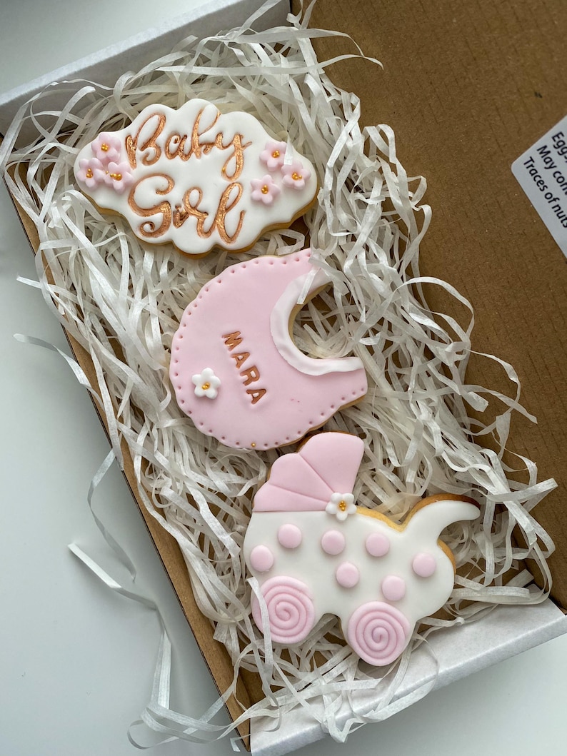 May include: A gift box filled with decorative cookies for a baby girl. The cookies include a cloud-shaped cookie with "Baby Girl" in gold script, a bib-shaped cookie, and a stroller-shaped cookie, all in pink and white.