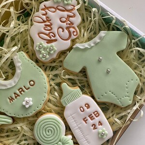 May include: A box of decorated baby-themed cookies. The cookies are in the shape of a bib, onesie, baby bottle, and a cookie with the text "Baby Girl". The cookies are decorated with green and white icing and silver sprinkles.