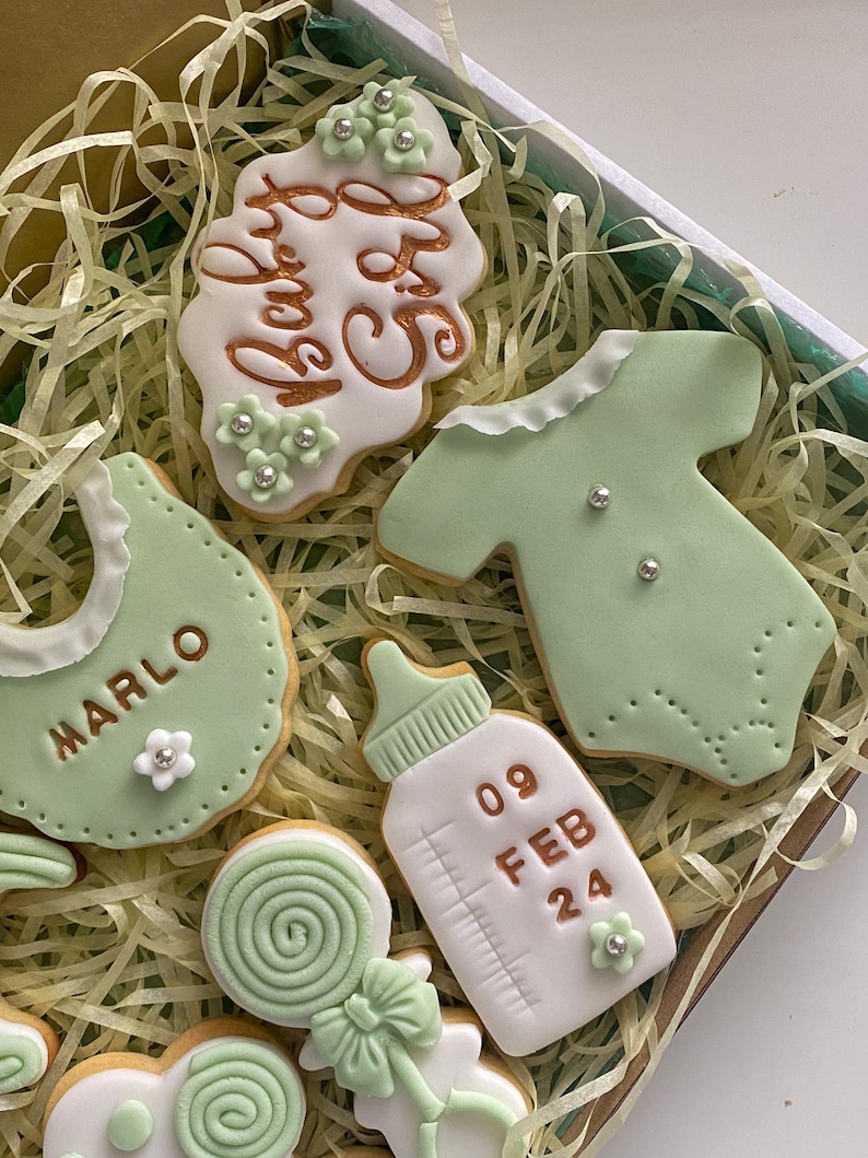May include: A box of decorated baby-themed cookies. The cookies are in the shape of a bib, onesie, baby bottle, and a cookie with the text "Baby Girl". The cookies are decorated with green and white icing and silver sprinkles.