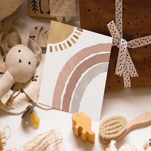 May include: A baby gift set with a variety of items, including a plush bunny rattle, a pacifier, a wooden dinosaur toy, a hair brush, a baby bib, and a card with a watercolor rainbow and sun design.
