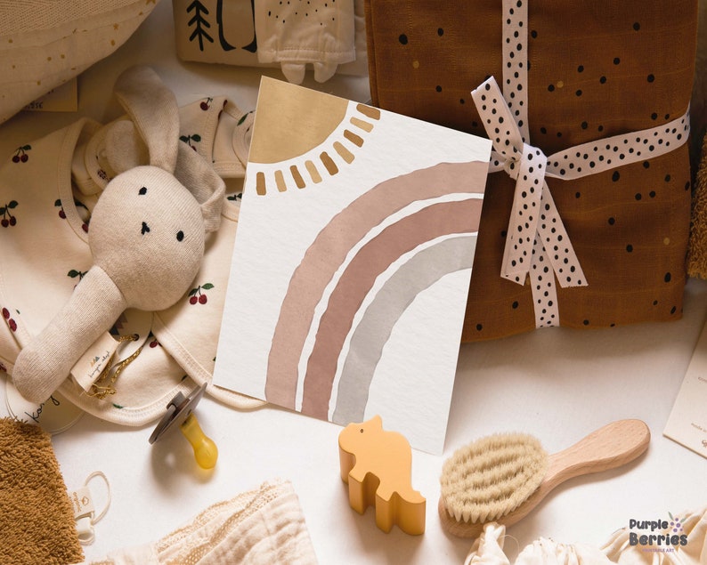May include: A baby gift set with a variety of items, including a plush bunny rattle, a pacifier, a wooden dinosaur toy, a hair brush, a baby bib, and a card with a watercolor rainbow and sun design.