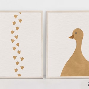 Puede incluir: Dos impresiones enmarcadas con un diseño minimalista. La impresión de la izquierda presenta una serie de huellas de patas de pato marrones en una línea vertical. La impresión de la derecha presenta una silueta de pato marrón.