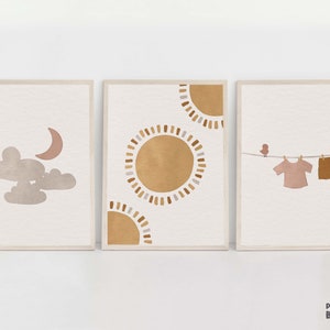 Puede incluir: Tres impresiones de arte minimalistas con fondo beige. La impresión de la izquierda presenta una nube gris y una luna creciente rosa. La impresión del centro presenta un gran sol marrón con un anillo blanco y marrón alrededor. La impresión de la derecha presenta un pequeño pájaro marrón y dos prendas marrones colgadas de una línea.