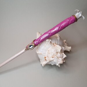 Könnte beinhalten: Ein handgefertigter Zauberstab mit einem hellen Holzschaft und einem leuchtend rosa, gesteppten Griff. Der Zauberstab hat einen dekorativen Schmuckstein und eine silberfarbene Spitze. Der Zauberstab ist etwa 30 cm lang.