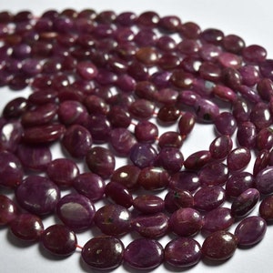 Op de afbeelding: Een streng dieprode robijn edelsteen kralen, elke kraal is ovaalvormig en gepolijst tot een gladde afwerking. De kralen zijn geregen op een dunne rode draad.